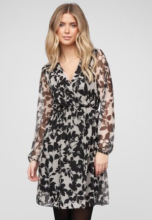 Blonde Frau, die ein schwarz-weißes Wickelkleid mit floralem Muster und langen, durchsichtigen Ärmeln trägt, vor einem schlichten hellen Hintergrund stehend.