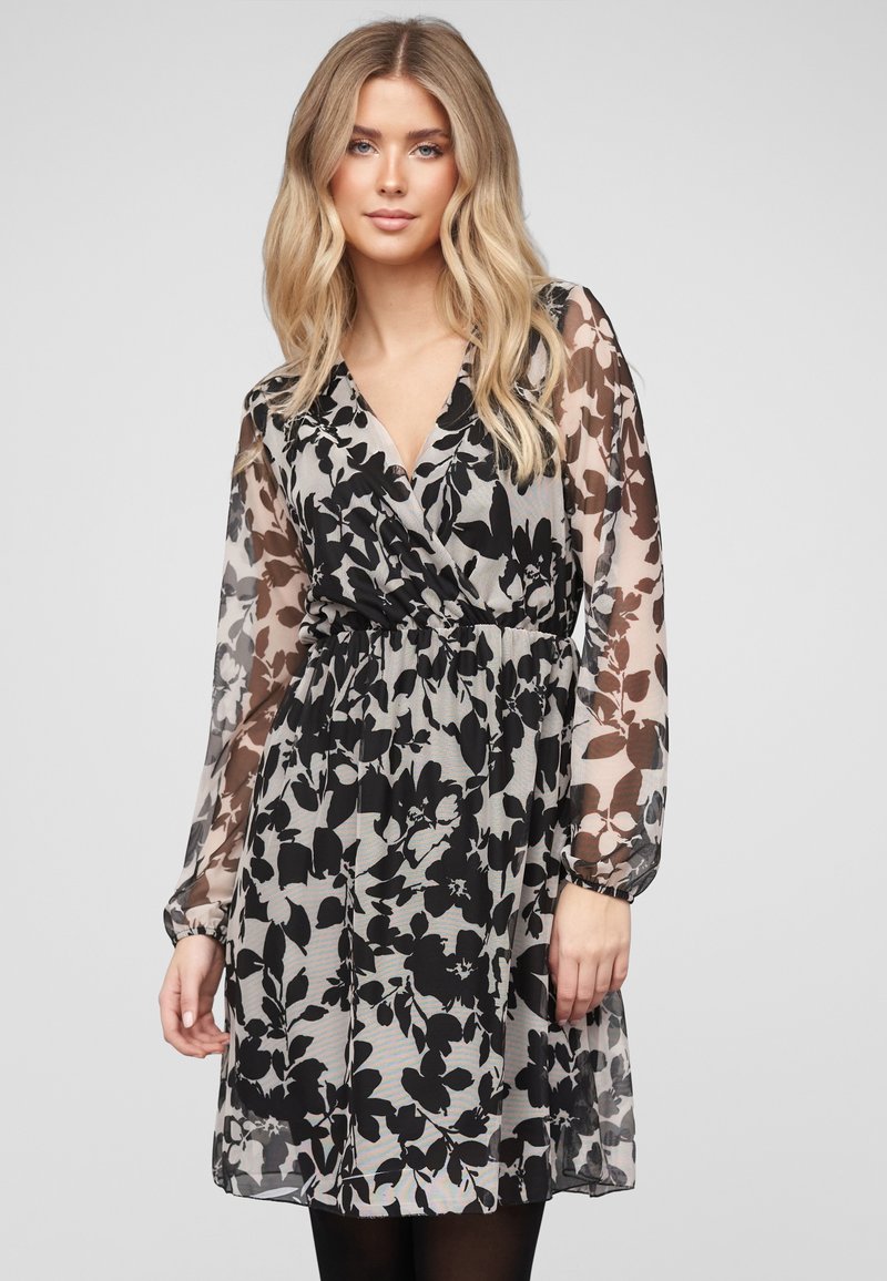 Lyshåret kvinde iført en sort og hvid blomstret slå-om-kjole med lange lette ærmer, stående foran en ensfarvet lys baggrund.