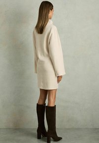 Rochie tricotată supradimensionată de culoare crem, cu mâneci lungi, guler înalt și fante laterale, asortată cu botine maro închis până la genunchi.