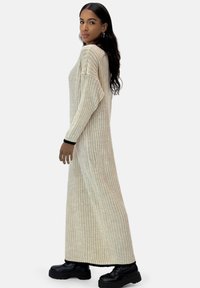 Langes, geripptes Strickkleid in hellbeige mit schwarzen Akzenten an den Handgelenken. Bietet einen lässigen Schnitt und einen Rundhalsausschnitt, kombiniert mit klobigen schwarzen Stiefeln.