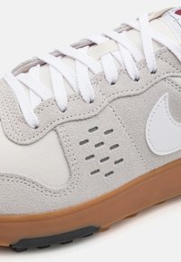 Sneaker grigio chiaro con tomaia in suede e tessuto, lacci bianchi, dettagli perforati e suola in gomma naturale. Logo Nike presente sul lato.