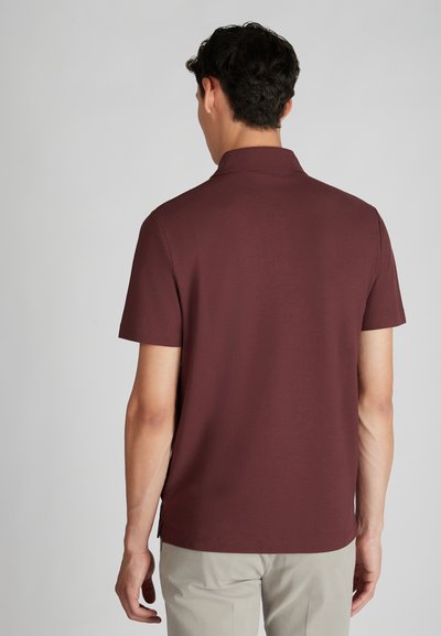 Man die wegkijkt, gekleed in een kortärmige bordeauxrode poloshirt en lichtbeige broek tegen een effen lichte achtergrond.