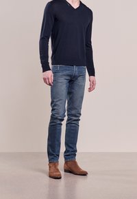 Mann in dunkelblauem Pullover, blauen Jeans und braunen Wildleder-Ankle-Boots steht vor einem beigen Hintergrund.