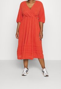 Robe rouge, style portefeuille avec des manches bouffantes trois-quarts, détail de ourlet plissé, fabriquée en tissu léger. Associée à des baskets blanches et grises.