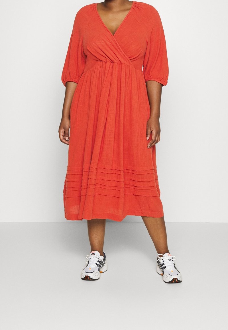 Robe rouge, style portefeuille avec des manches bouffantes trois-quarts, détail de ourlet plissé, fabriquée en tissu léger. Associée à des baskets blanches et grises.