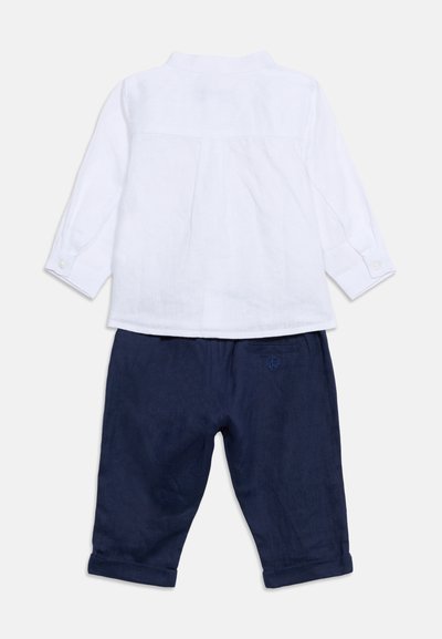 Λευκό παιδικό πουκάμισο με μακριά μανίκια και κουμπωτές μανσέτες, συνδυασμένο με σκούρο μπλε παντελόνι με ρεβέρ, εμφανίζεται από πίσω σε απλό φόντο.