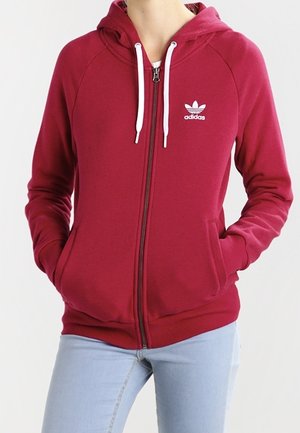 Osoba ubrana w czerwoną bluzę z kapturem i suwakiem Adidas z białymi sznurkami oraz jasnoniebieskie dżinsy, dłonie w kieszeniach bluzy, stojąca na tle jednolitego tła.