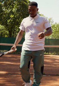 Tennisser in een wit polo-shirt met een groen Nike-logo en groene broek met zwarte accenten, die een tennisracket vasthoudt op een gravelbaan.