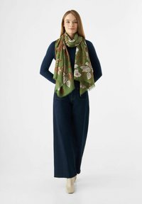 Écharpe verte avec imprimé floral en marron et blanc, portée sur un haut marine. Le modèle est associé à un jean taille haute à jambe large et des bottes crème.