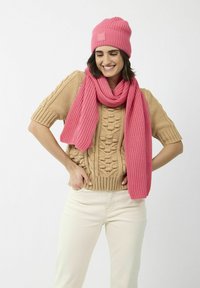 Pull en laine beige avec un motif texturé, associé à un bonnet rose côtelé et une écharpe assortie. Matériaux doux et chauds. Fond clair.