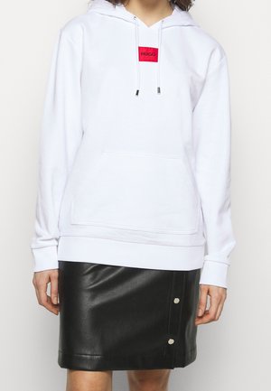 Person wearing a white hoodie with a red "HUGO" label and a black leather skirt with two visible snap buttons.