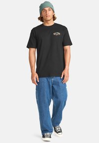 Black cotton t-shirt with a colorful logo on the left chest, paired with loose blue denim jeans and black sneakers with white soles.