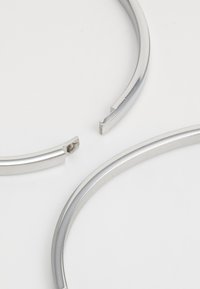 Pulseras de metal plateado con un acabado liso y brillante. Una pulsera muestra una ruptura con un detalle interior visible. Diseño simple y curvado.