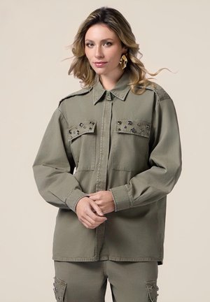 Camicia utilitaria verde oliva in cotone, con due tasche sul petto con decorazioni nere, chiusura con bottoni e maniche lunghe.