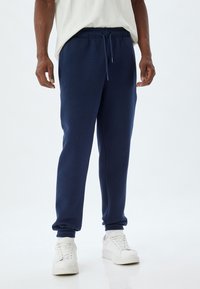 Pantalones de chándal azul marino con puños elásticos, cintura con cordón y bolsillos laterales, hechos de material suave, combinados con zapatillas blancas.