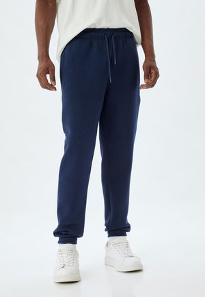 Mørkeblå sweatpants med elastiske manchetter, snoretræk i taljen og sidelommer, lavet af blødt materiale, parret med hvide sneakers.