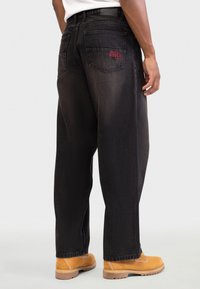 Crne traperice s labavim krojem, imaju dva stražnja džepa; jedan džep ima crvenu vezanu riječ "RODEO". Nošene uz smeđu obuću.