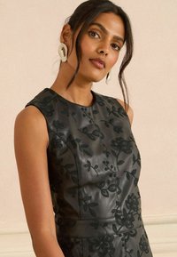 Rochie din piele neagră cu broderie florală, design fără mâneci, guler înalt și o siluetă ajustată cu detalii vizibile ale cusăturilor.