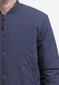 Námorníkový prošívaný bomber s ribovým límcem, zipovým uzávěrem, bočními kapsami a texturou látky s diamantovým vzorem.