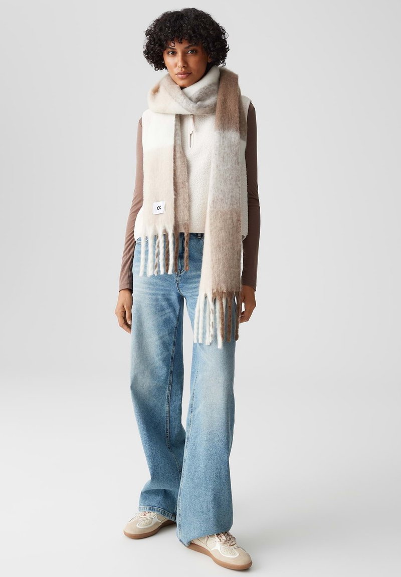 Sciarpa lavorata a maglia con strisce beige e crema, con estremità frangiate e un piccolo logo ricamato. Indossata con un top senza maniche e jeans a gamba larga.