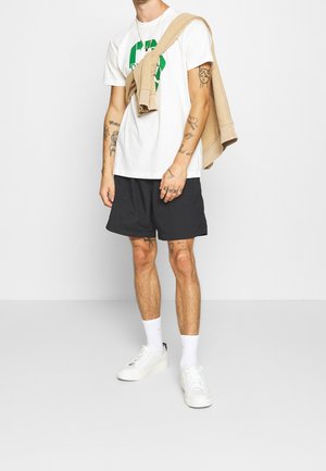 Hombre con camiseta blanca con estampado verde, pantalones cortos negros, zapatillas blancas y sudadera beige sobre los hombros, mostrando tatuajes en el brazo y la pierna.