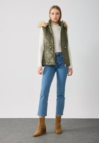 Quilted olive green vest with faux fur hood, zip closure, and side pockets, paired with blue straight-leg jeans and tan heeled ankle boots.