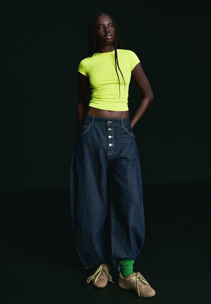 Femme aux longues tresses portant un haut court jaune fluo, un jean large bleu foncé avec des boutons, des chaussettes vertes et des baskets beiges sur fond noir.