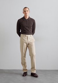 Brun zip-hals sweater, cremede bukser, polerede brune sko; modellen står mod en lys grå baggrund. Enkel, formel outfit udseende.