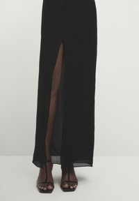 Schwarzes Maxikleid mit einem Oberschenkelspalt, aus leichtem Stoff gefertigt. Es hat eine glatte Textur und wird mit schwarzen Sandalen mit quadratischer Zehenform kombiniert.