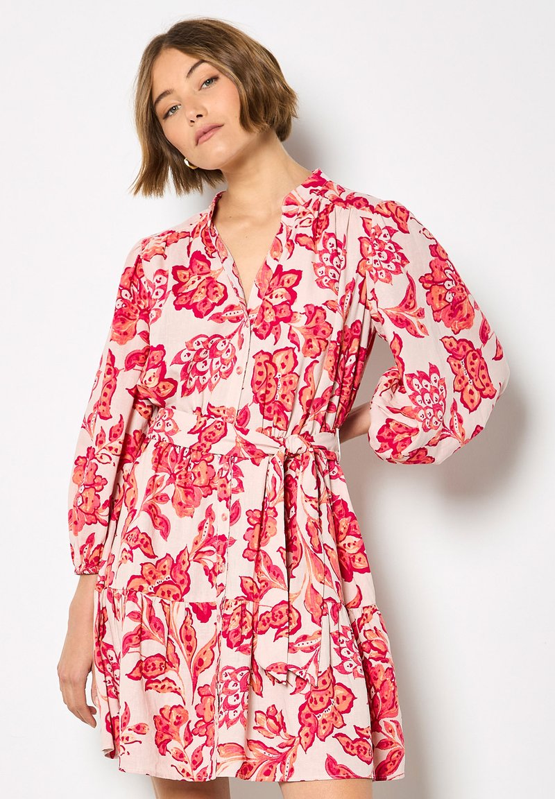 Robe à imprimé floral avec des fleurs roses et rouges sur un fond clair. Elle présente un devant boutonné, une taille à nouer et de longues manches bouffantes.