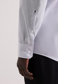 Camicia bianca a maniche lunghe con un piccolo motivo a quadretti blu, polsini con bottoni e dettaglio di fiore nero ricamato sulla manica.