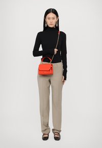 Rød læderhåndtaske med håndtag og kædestrop, der har en klaplukning. Modellen bærer en sort turtleneck og beige bukser.