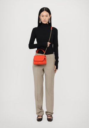 Sac à main en cuir rouge avec une poignée supérieure et une chaîne, doté d'une fermeture à rabat. Le modèle porte un col roulé noir et un pantalon beige.