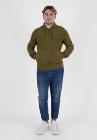 Felpa con zip color verde oliva, dotata di cordini, abbinata a jeans blu e sneakers bianche. Il materiale sembra morbido con una finitura testurizzata.