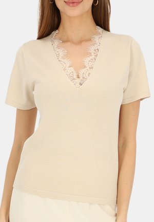 Kobieta w beżowej bluzce z krótkim rękawem, z dekoltem w serek ozdobionym beżową koronką i małymi guzikami.