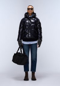 Giacca a vento nera lucida con cappuccio, guanti grigi senza dita, camicia bianca, jeans blu e stivali alla caviglia marrone scuro; tiene una borsa nera.