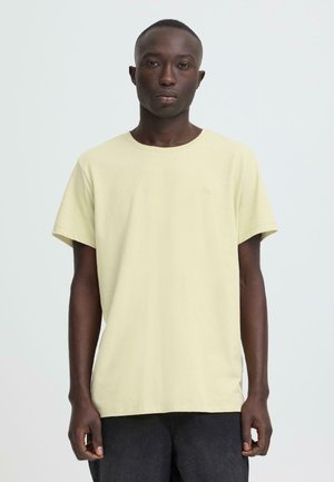 Donkere man, staand en recht vooruit kijkend, met een effen lichtgele korte mouw t-shirt en donkere broek tegen een witte achtergrond.