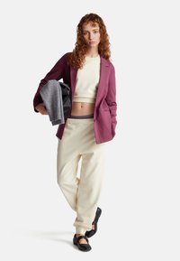 Blazer bordeaux, maglione corto beige, pantaloni da tuta color avorio e scarpe nere. Tieni un maglione grigio. Capelli ricci e sfondo neutro.