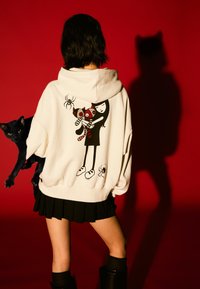 Biała bluza z kapturem z grafiką dziewczyny z czerwono-czarnymi pasiastymi kotami, uzupełniona czarną plisowaną spódnicą i czarnym kotem. Czerwone tło.