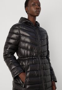 Manteau en duvet noir avec capuche, présentant des sections matelassées horizontales, une taille cintrée, une fermeture éclair à l'avant et des poches latérales. Fini brillant et léger.