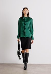 Grüne Satinbluse mit Rüschendetails auf der Vorderseite, kombiniert mit einem schwarzen Bleistiftrock und knielangen schwarzen Stiefeln. Das Model trägt eine Handtasche.