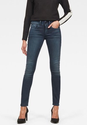 Jeans ajustados en denim azul oscuro, con un ligero desgarro, que presentan un diseño de cinco bolsillos y un sutil detallado de costura. Los usuarios los combinan con tacones negros.