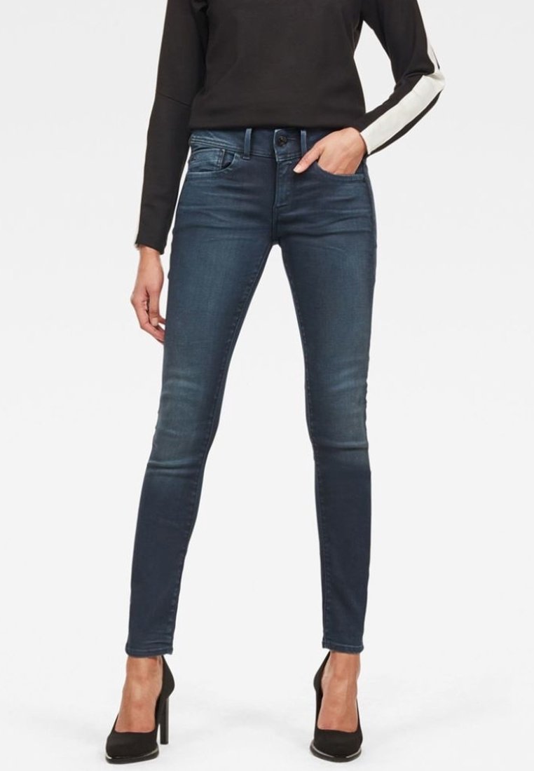Jeans ajustados en denim azul oscuro, con un ligero desgarro, que presentan un diseño de cinco bolsillos y un sutil detallado de costura. Los usuarios los combinan con tacones negros.