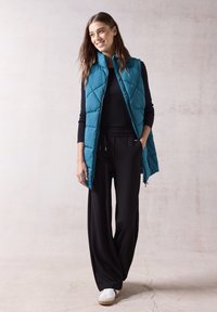 Turkos pufferväst med diamantprickning, i kombination med svarta vida byxor. Modellen bär vita sneakers och står mot en neutral bakgrund.