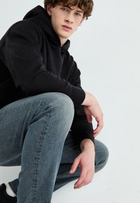 Fiatal, barna, göndör hajú férfi fekete kapucnis pulóverben és kék farmerben guggol egy egyszerű fehér háttér előtt.