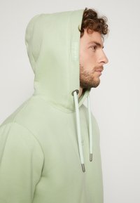 Lichtgroene hoodie, zachte stof, kangoeroezak, verstelbare koordjes met metalen uiteinden, klein logo op de borst, relaxte pasvorm.