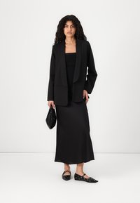 Fekete szabott blézer egy testhez simuló fekete ruhán, elegáns fekete lapos cipővel és egy kis fekete kézitáskával. Sima anyag és minimalista dizájn.