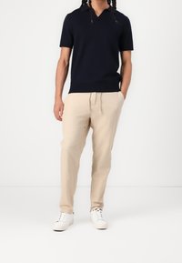 Marinblå kortärmad polo med V-ringning, kombinerad med ljusbeiga dragsko-byxor och vita sneakers mot en enfärgad bakgrund.