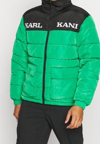 Giacca a vento verde e nera con colletto alto, chiusura con zip e due tasche frontali; presenta un testo logo bianco sul petto.