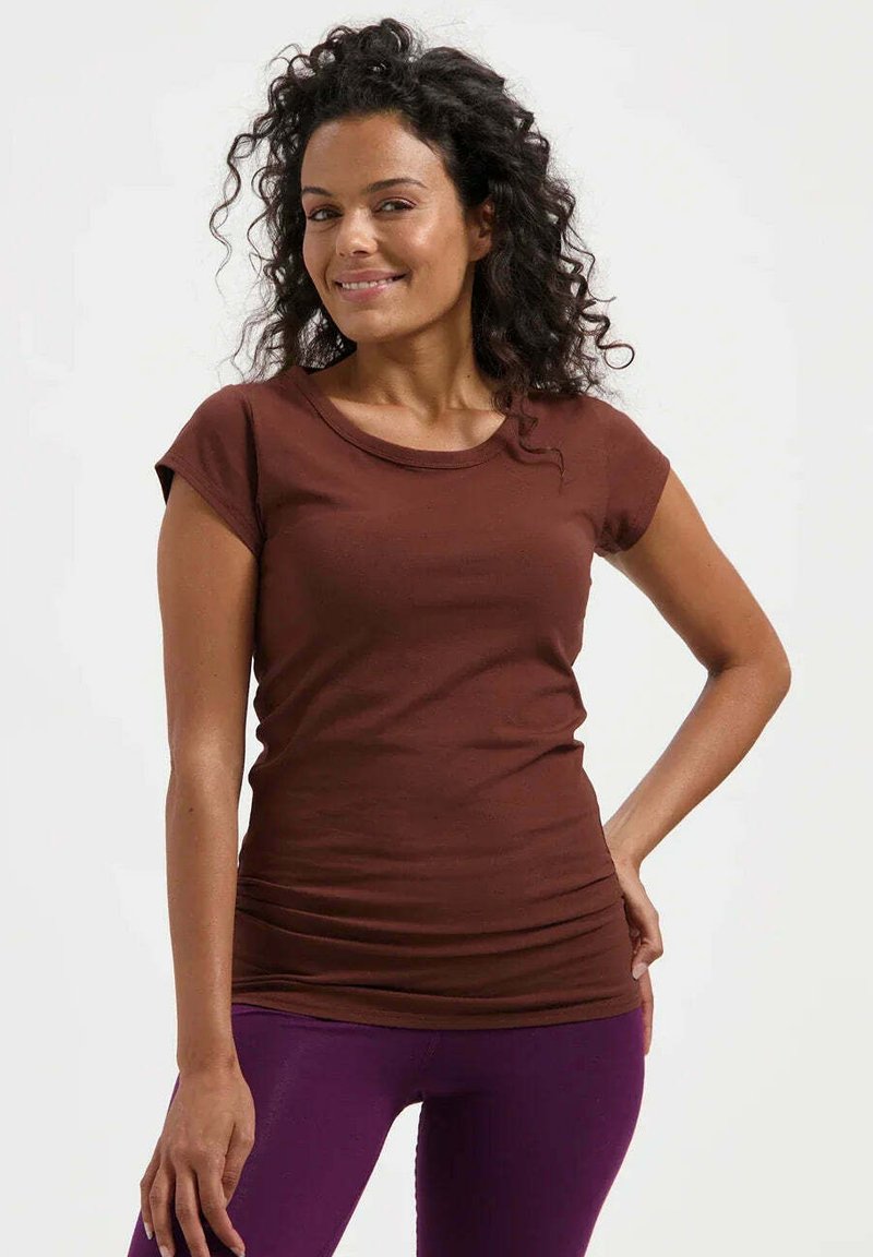 Femme souriante aux cheveux bouclés, portant une chemise marron à manches courtes et un pantalon violet, debout avec une main sur la hanche.